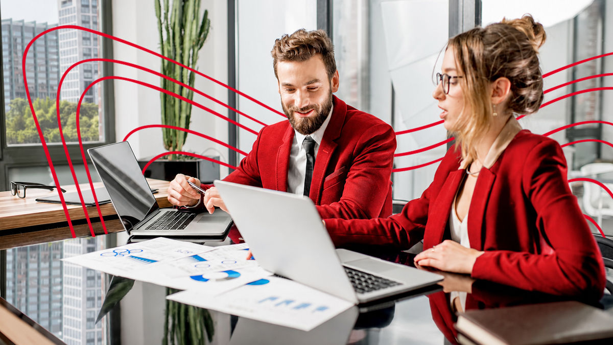A man and woman wearing matching red blazers sit in an office building at a glass table. Laptops and print-outs of graphs and charts clutter the table. The man and woman are deep at work but they both appear happy with their collaboration and smile. 