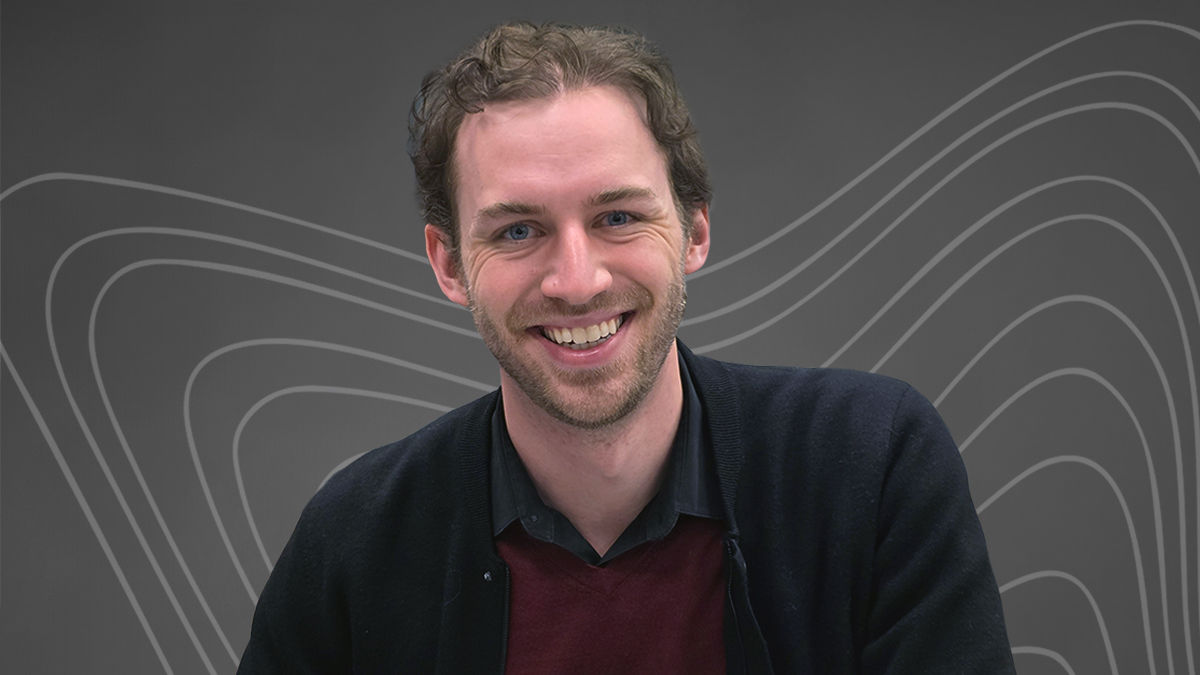 UC online MBA graduate Mike Carovillano smiles in front of a gray background. He is white, has short curly brown hair, and wears a black cardigan over a burgundy collared shirt.