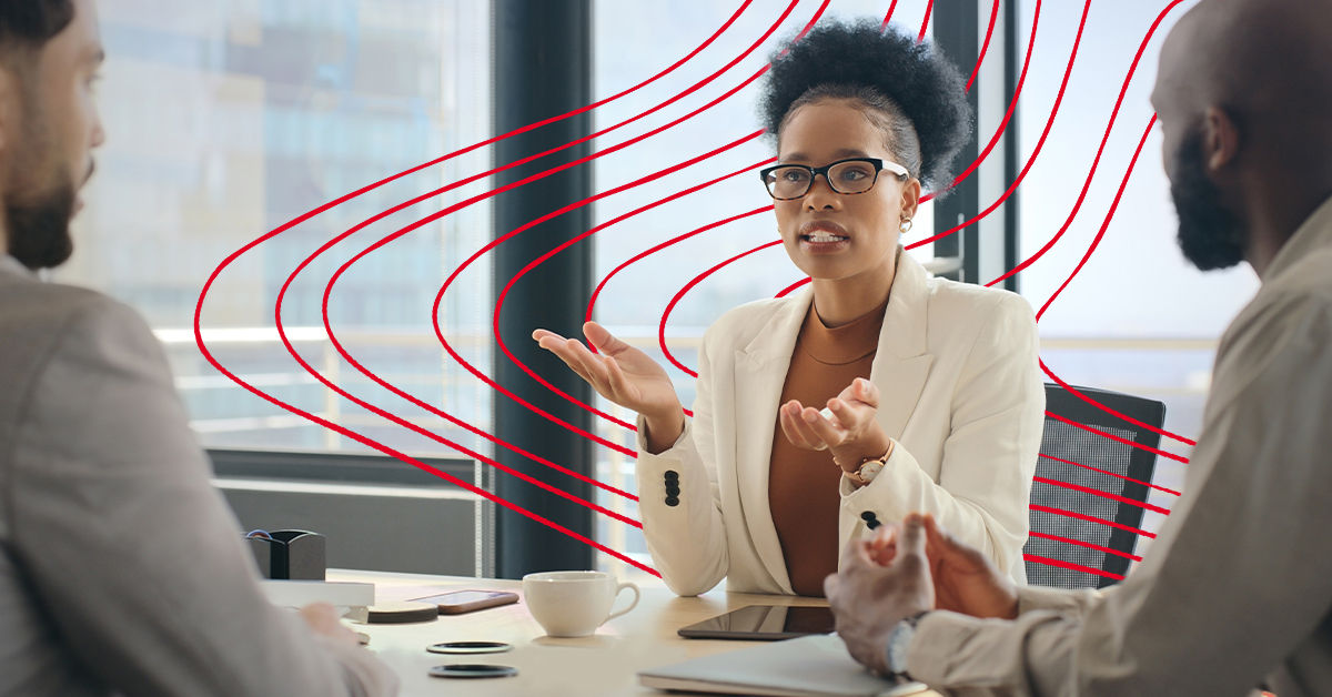A woman sits talking at a desk in an office. She wears a white blazer and brown glasses, and her hands are raised as she speaks. Two men wearing business clothes sit across from her at the table. 