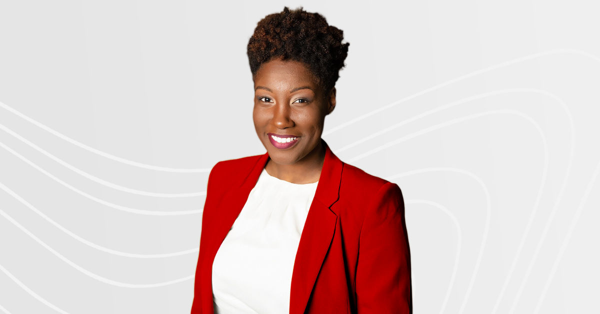 UC professor Dr. Monyae Williamson-Gourley stands in front of a light gray background and smiles. She is black, has short curly dark hair, and wears a red blazer over a white blouse.