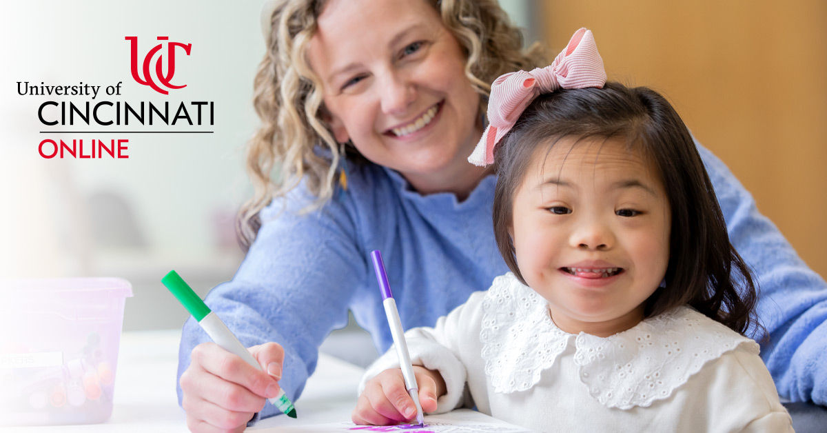 A female teacher with curly blonde hair sits at a table coloring with a female child with special needs. Both are smiling and holding markers. 