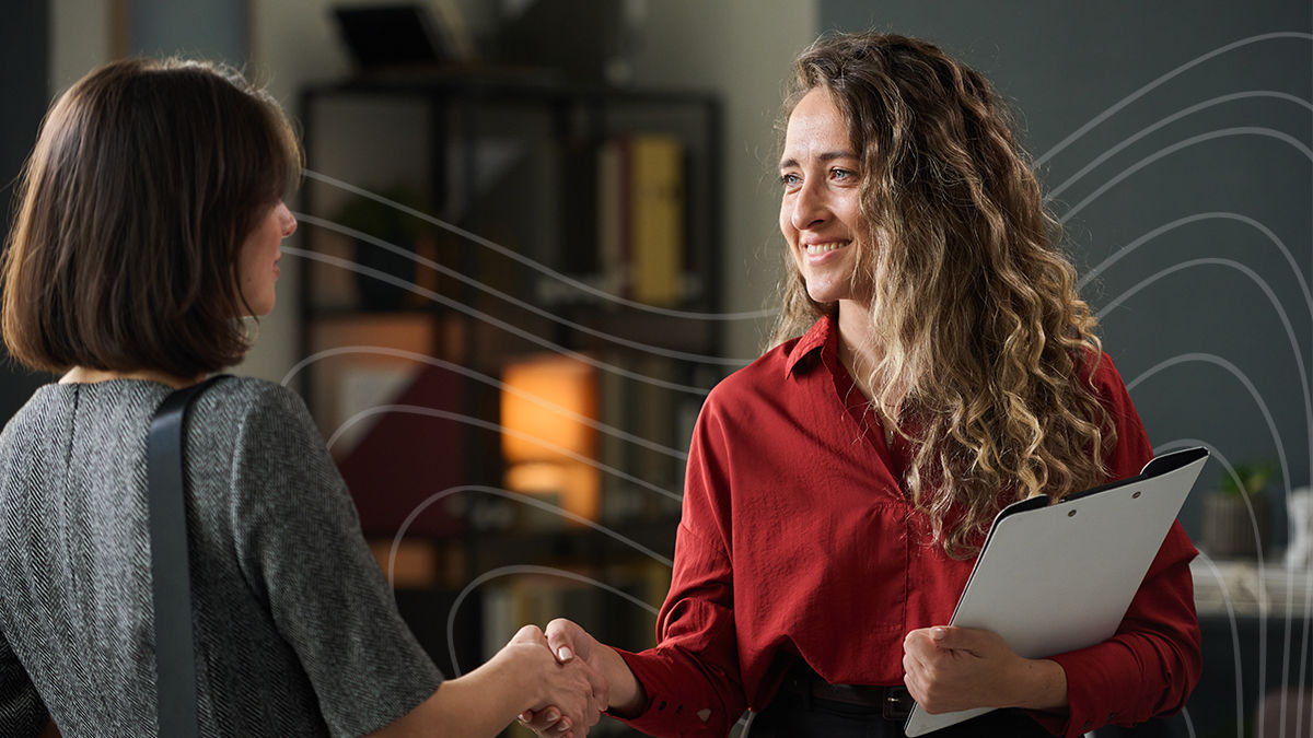 A woman in a red blouse holding a clipboard shakes hands with another woman in business attire. They are both in an office setting