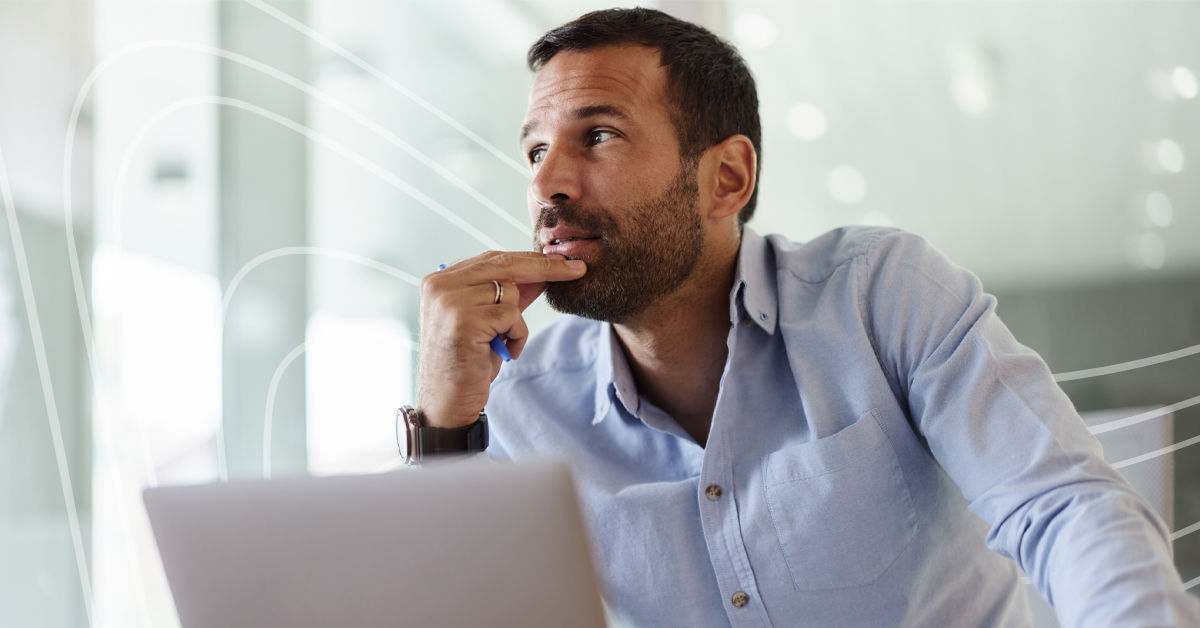 A man with dark hair and a beard sits in front of an open silver laptop. He wears a light blue button up shirt and has two fingers poised against his chin. He looks off to the side as though he’s deep in thought.