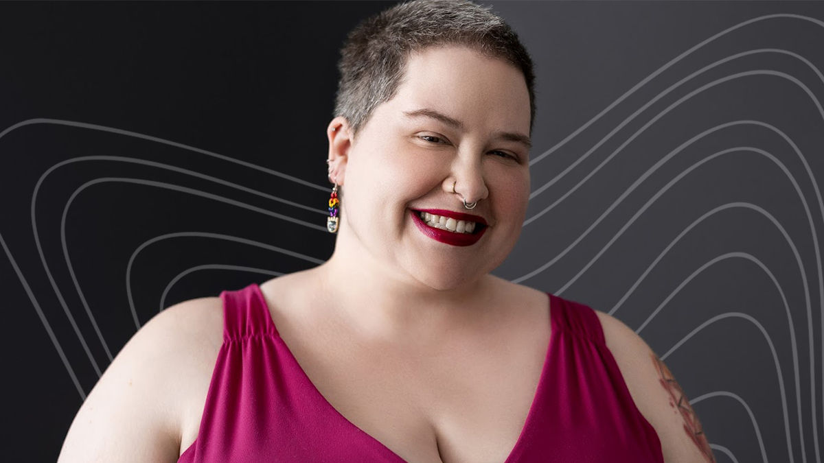 UC Online student Jeanne Delahunty wears a magenta short sleeved blouse and smiles. She has short brown hair and wears bright red lipstick and colorful rainbow earrings.