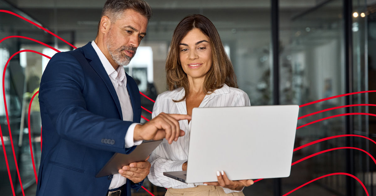 A woman in a white blouse holds her laptop screen towards a male colleague wearing a blue suit. He points at the screen. They are both in an office setting. 