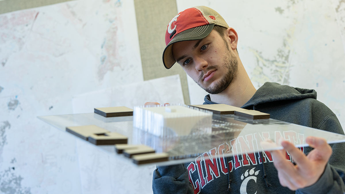 A male student in a University of Cincinnati hoodie and baseball cap holds a 3D model of an urban planning design. He stares at it with concentration. 