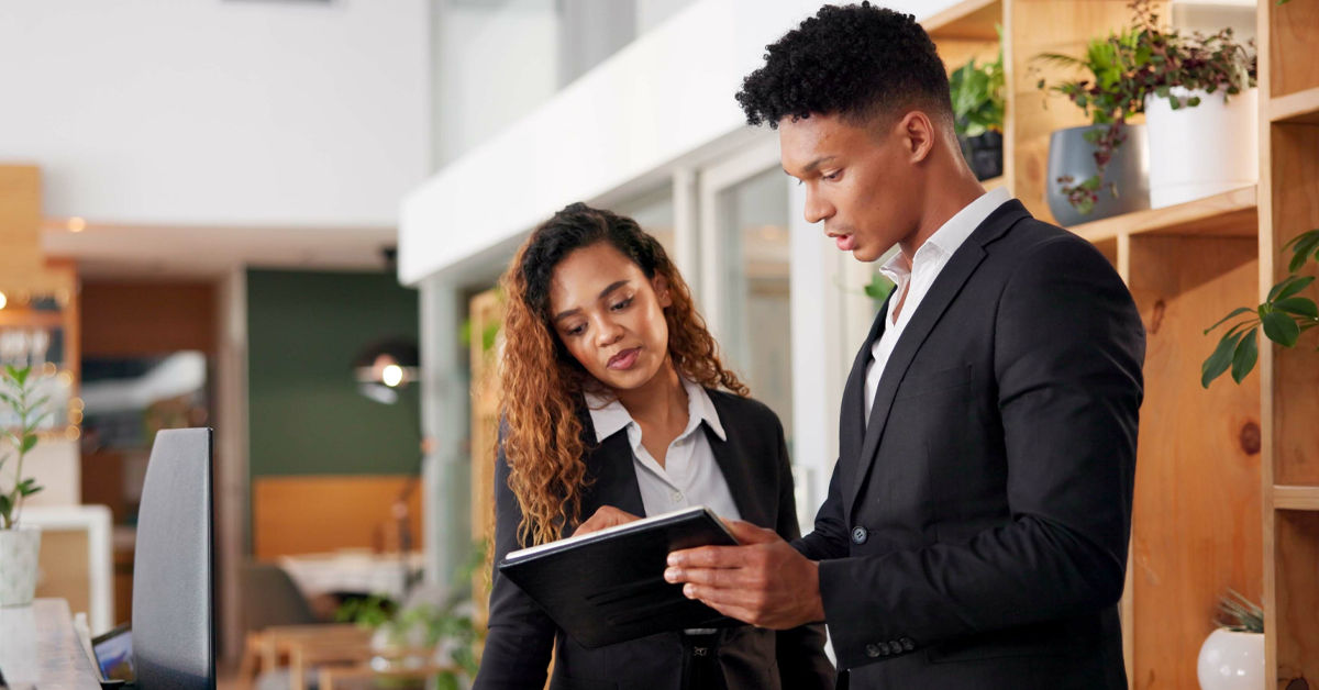 A man in a black business suit stands with a female colleague who is also wearing a business suit and white blouse. They both look at a tablet the man is holding, in an office setting. 