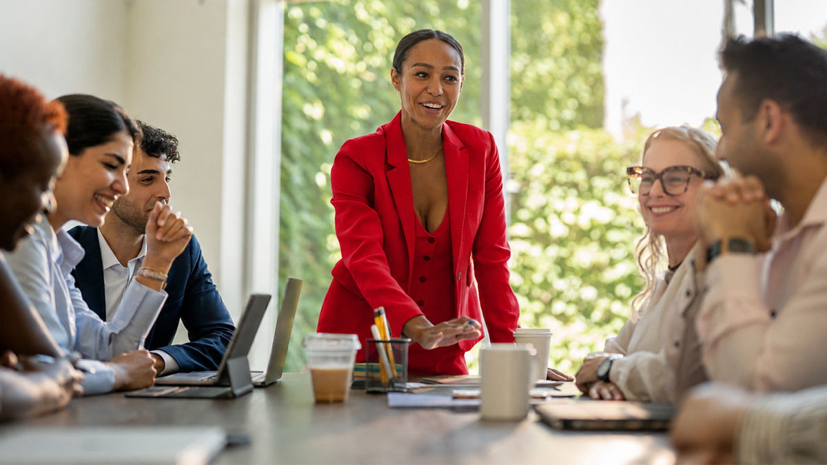 A woman stands at the head of a business meeting table wearing a red suit. She is smiling and speaking to one of her seated co-workers. Everyone in the room grins and looks happy. The table is cluttered with laptops and coffee cups. 