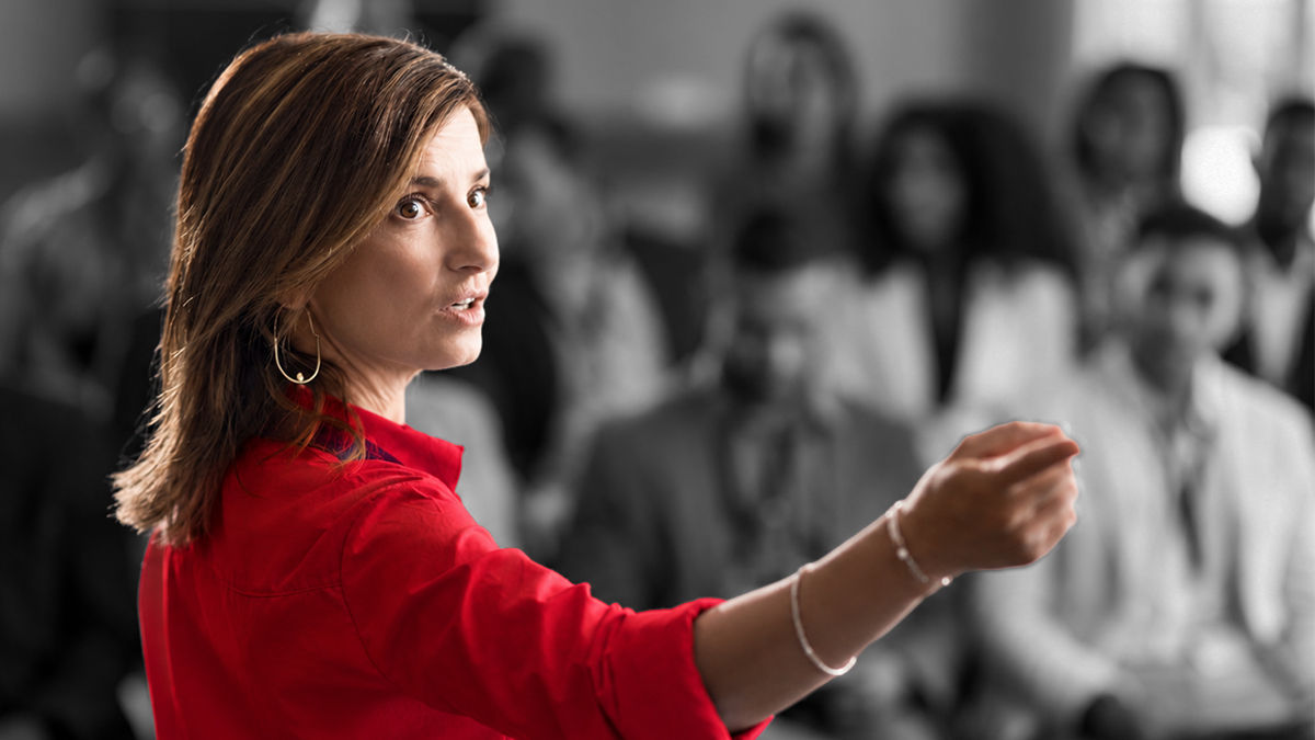 A woman in a red business shirt stands in front of a crowd, speaking. Her hand is pointed to a presentation screen offscreen. 