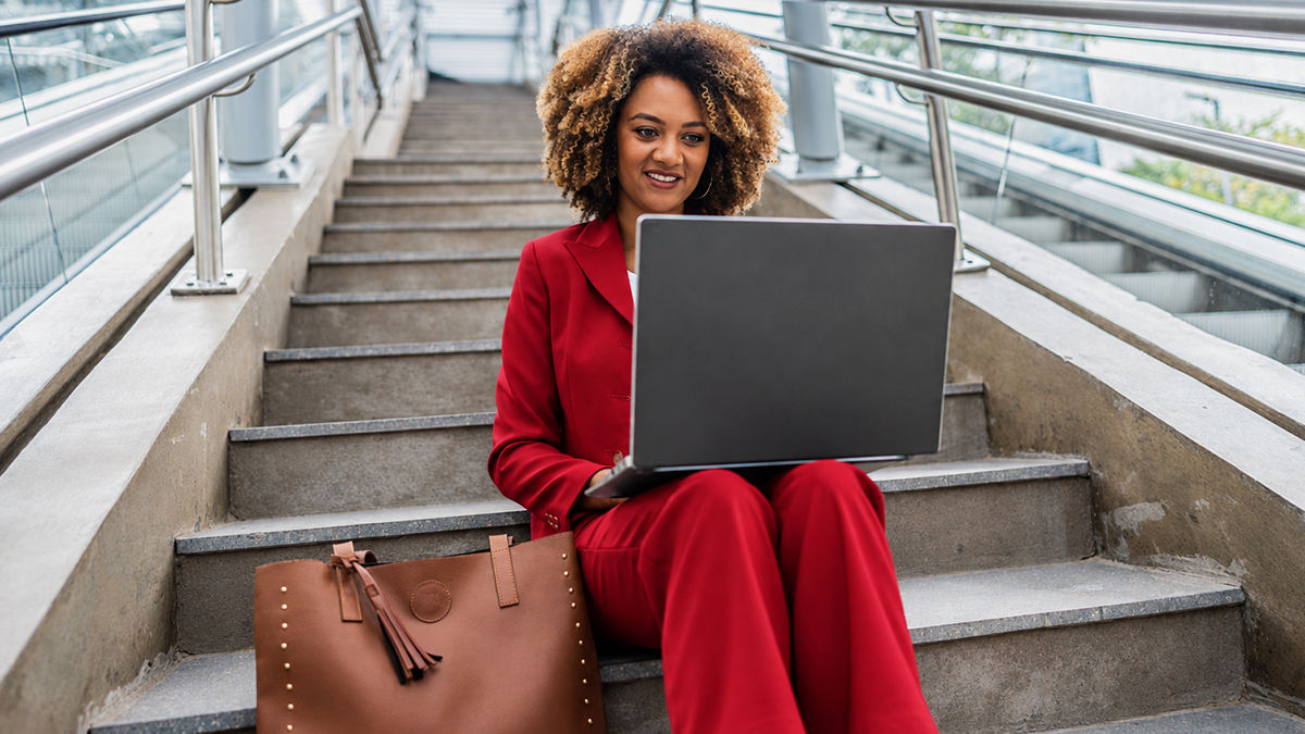 A woman in a red suit sits outdoors on concrete steps working on her laptop. She is smiling and looks engaged. 