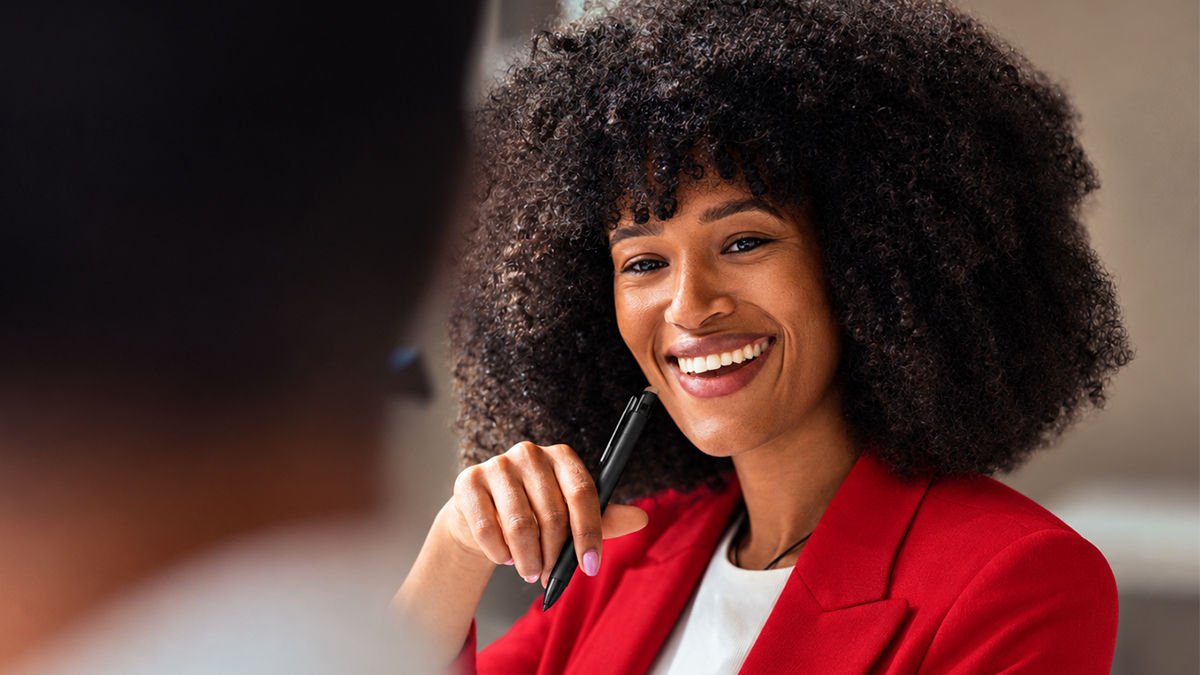 A woman in a red business suit smiles in an office setting while looking at a co-worker offscreen. She holds a pen close to her face as she thinks. 