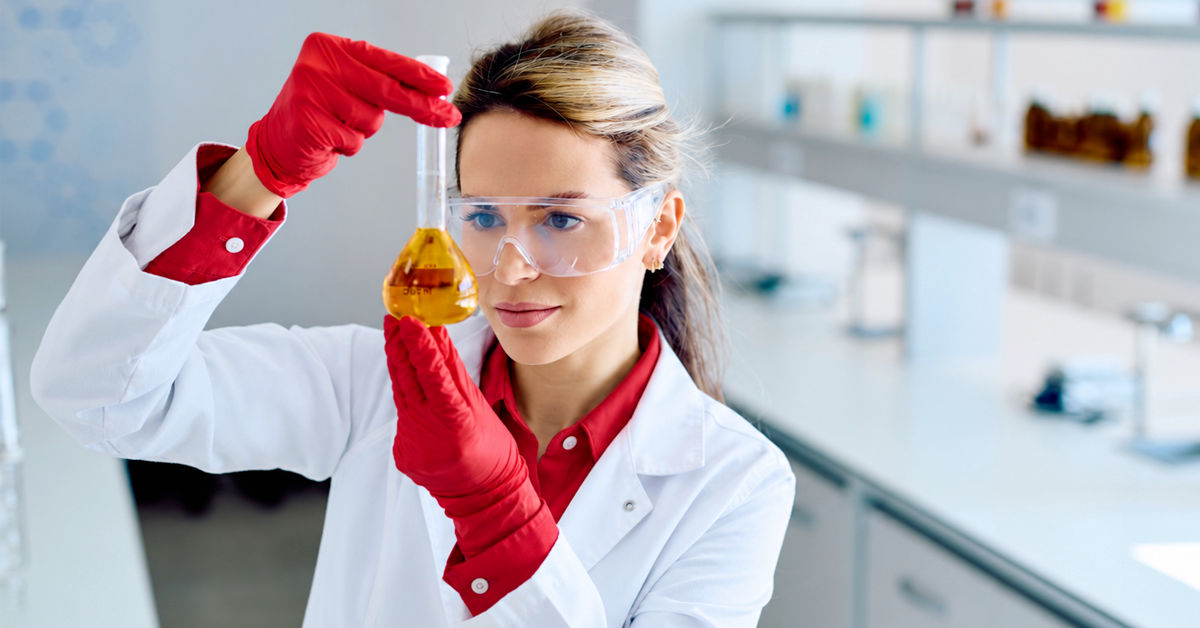 A woman in a laboratory setting wearing a white lab coat and goggles holds a glass beaker close to her face. It is filled with a yellow liquid. She looks intently inside. 