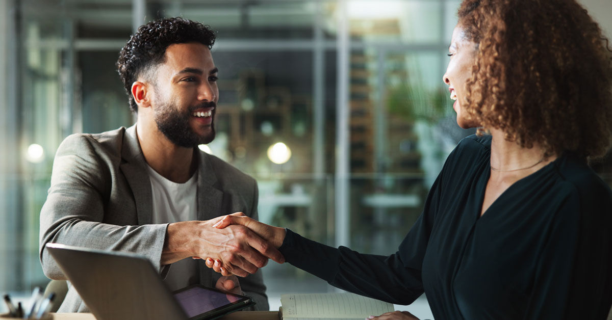 Man with dark brown hair wearing a gray business suit shaking hands with a female business partner with curly brown hair wearing a black button down shirt.