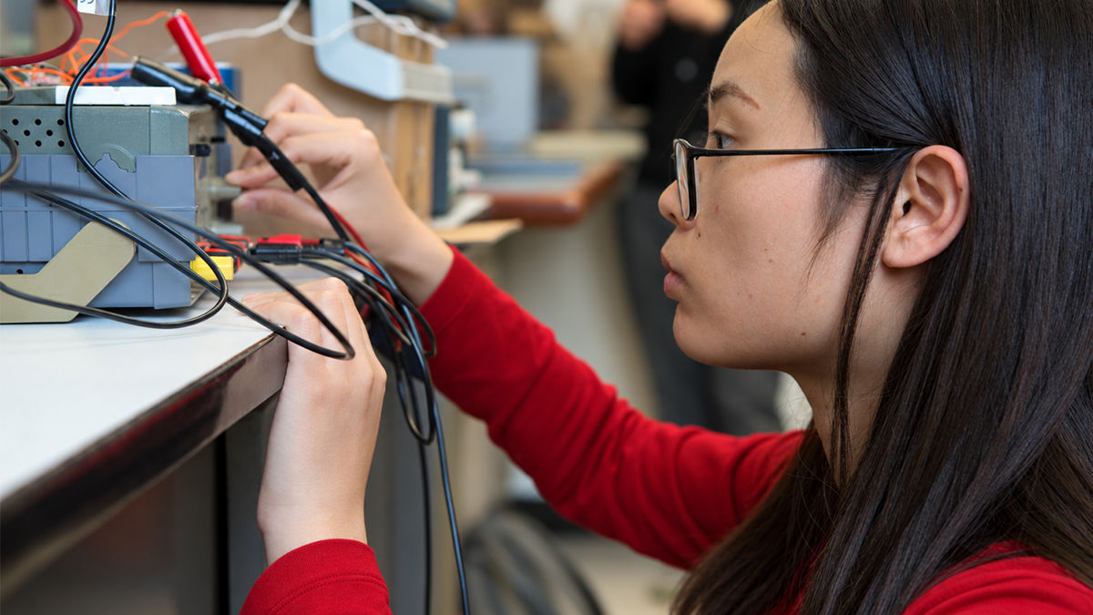 A woman in a red shirt and black glasses looks at a circuit system with extensive cords; she adjusts a dial and looks intently at the system. 