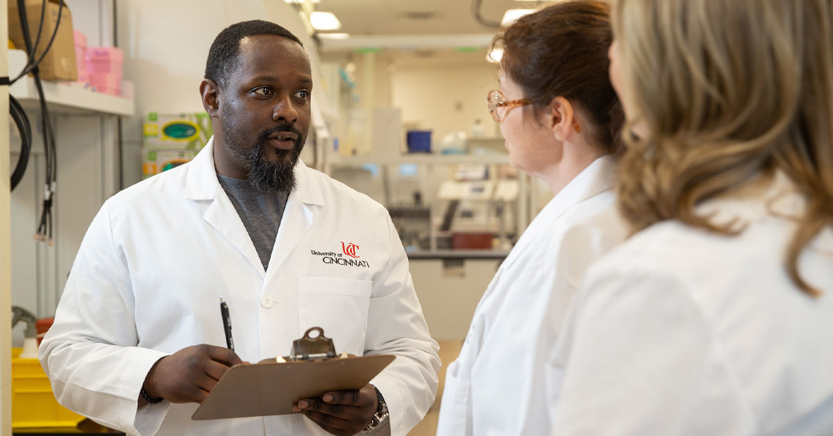 A man in a white University of Cincinnati lab coat holds a clip board and pen in a medical laboratory setting; he speaks to two female colleagues also wearing white lab coats. 