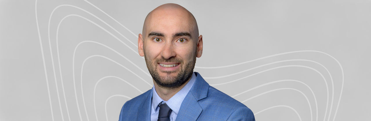 Dr. Rhys Axon wears a blue blazer and light blue collared shirt with a navy tie. He is bald, has a beard, and smiles in front of a light gray background.