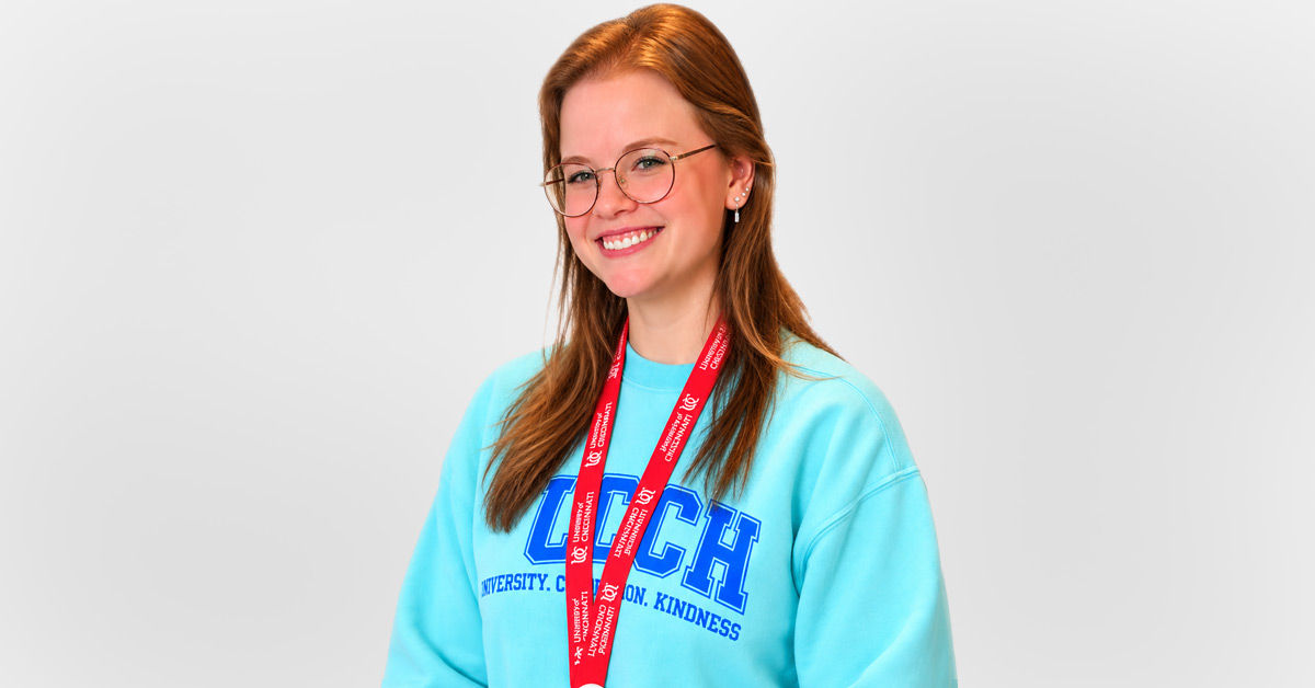Meghann Shalosky wears a bright blue longsleeve shirt and a red lanyard around her neck. She has red hair and glasses and smiles in front of a light gray background.