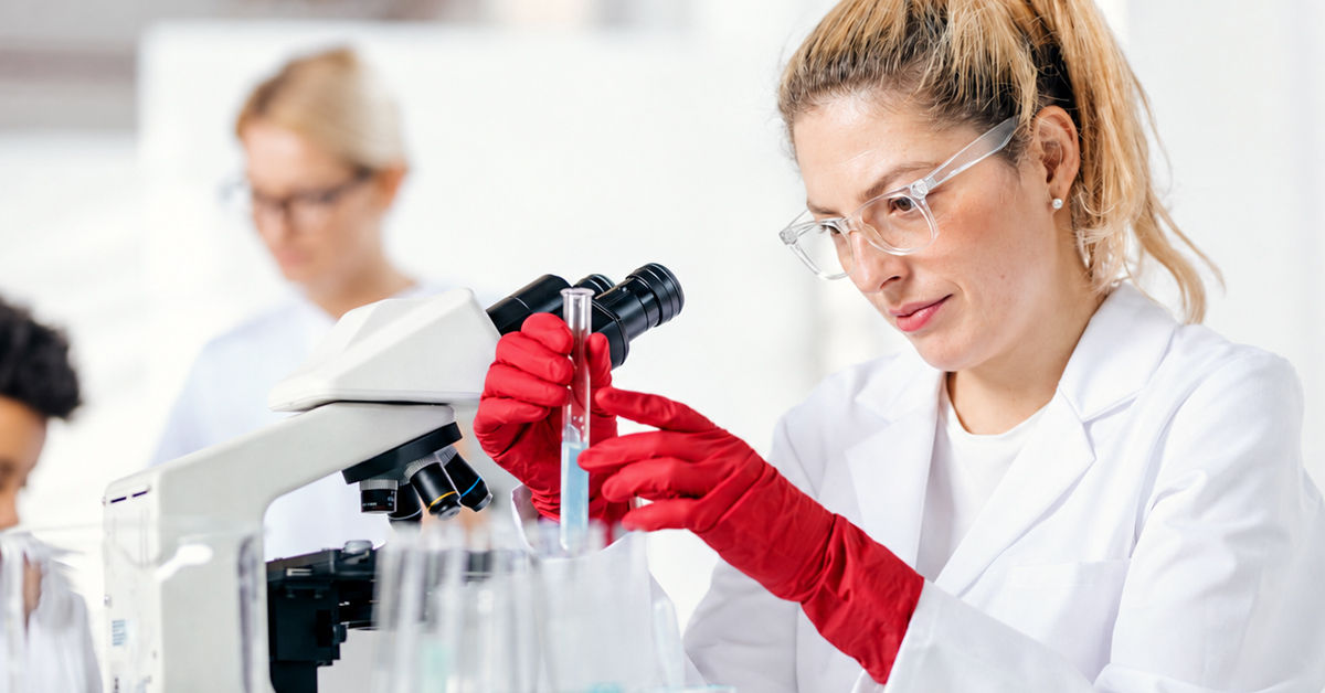 A woman in a white lab coat and red gloves stands holding a glass beaker beside a large microscope. She wears safety googles. 