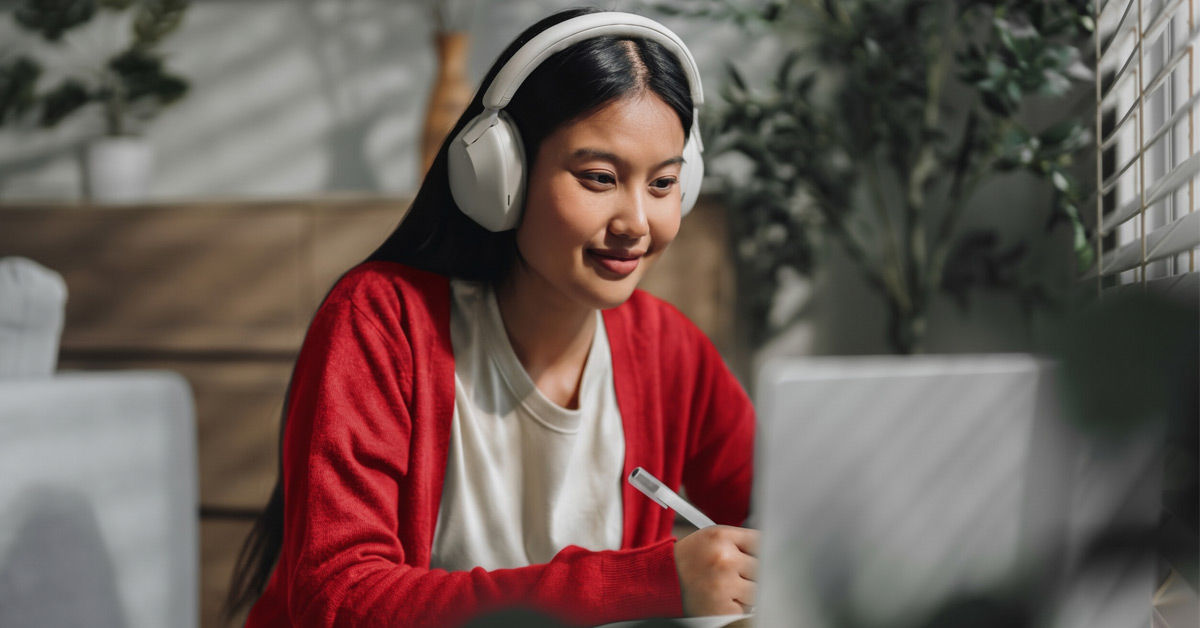 A girl with dark long hair wears a beige sirt and a red longsleeve cardigan. She sits in front of a grey laptop in a cafe or living room setting. She holds a pen and wears over-ear gray headphones.