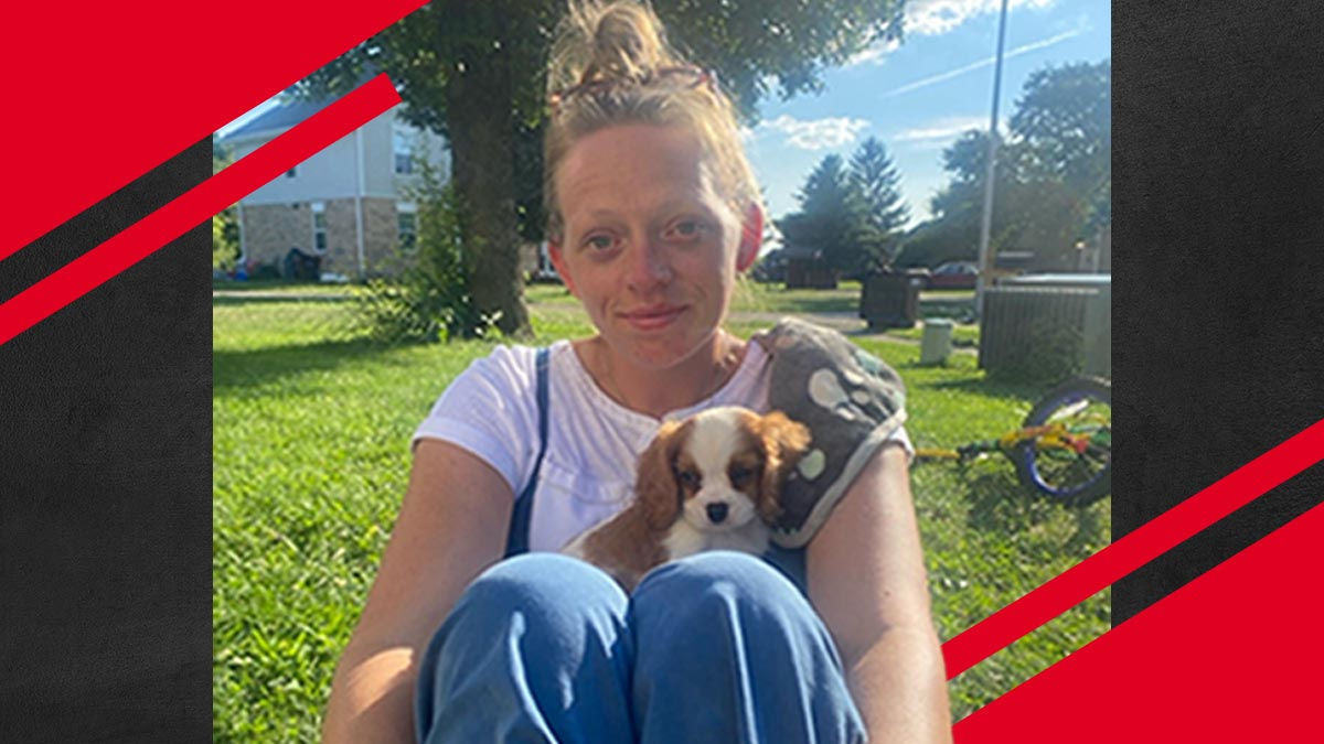 UC online student Maggie Brisker sits outside in a grassy backyard setting. She has light blond hair pulled into a high bun. She wears a white t-shirt, blue jeans, and holds a cocker-spaniel puppy in her lap.