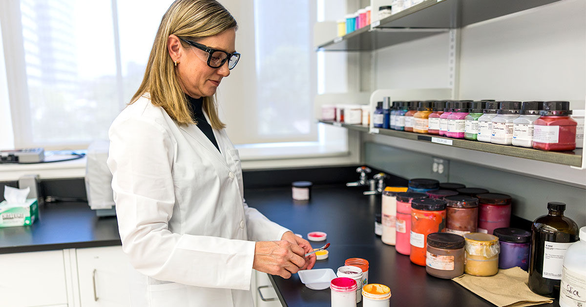 A woman in a laboratory setting wearing a white lab coat and goggles holds a spoon of a sample of cosmetic powder to be examined.