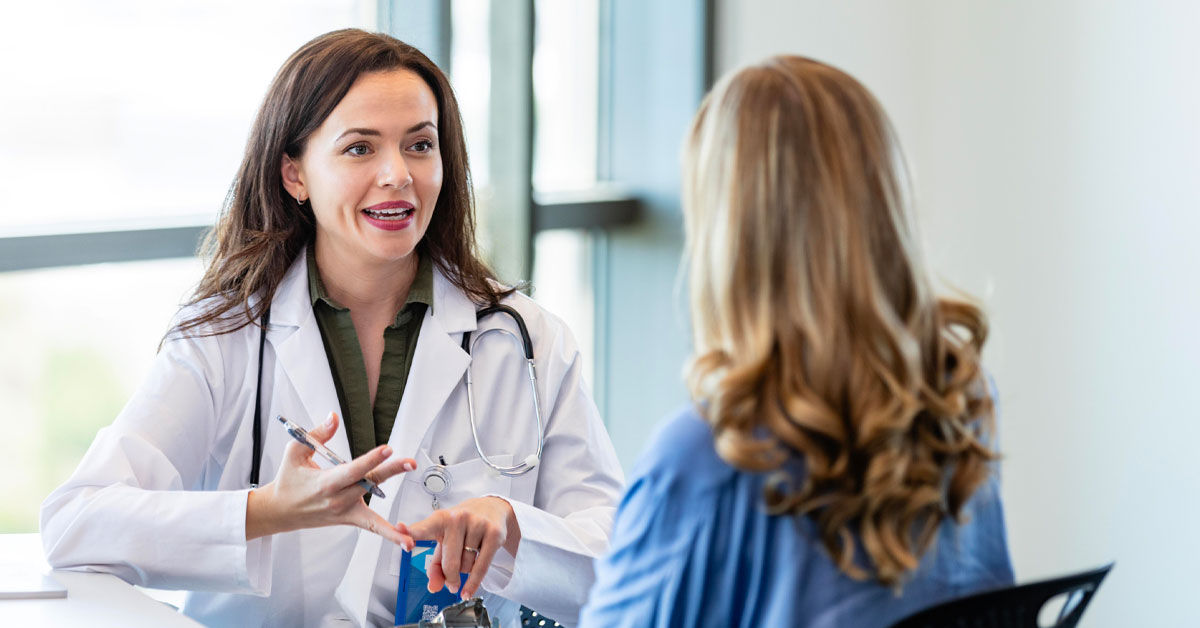 A female doctor wearing a white doctor's coat sits at a table and speaks to a female patient with long blonde hair. 