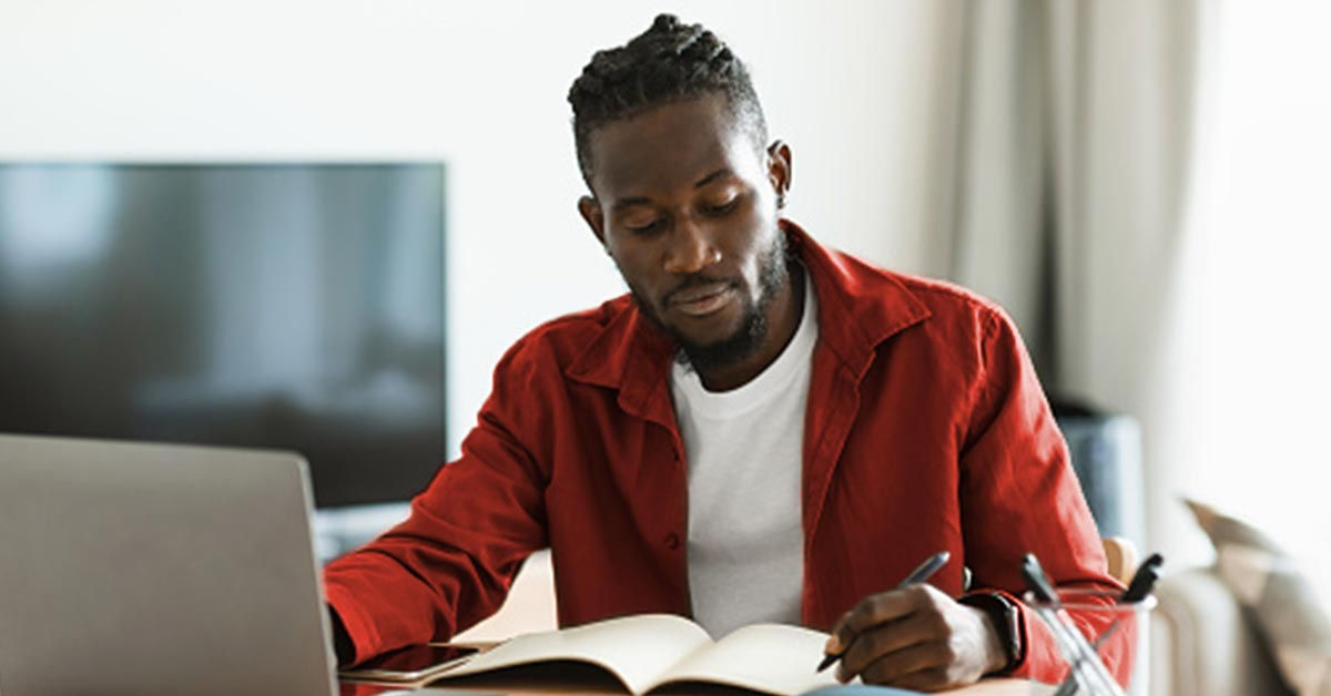 A black man sitting at a desk with his laptop open and a notebook open in front of him that he takes notes in.