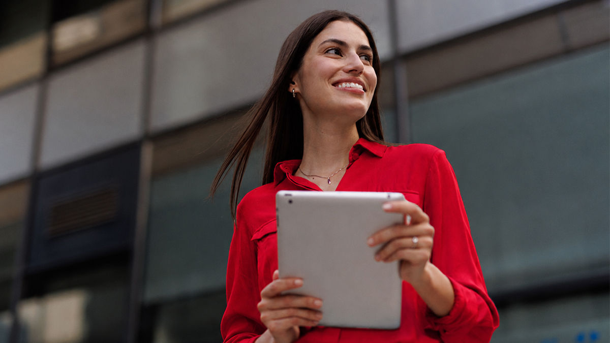 A woman with dark long straight wear wears a UC red long sleeve blouse. She stands outside and smiles in front of a building on campus. She holds a gray tablet in front of her.