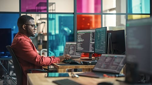 A software Engineer working on set of coding programs on multiple computer monitors.