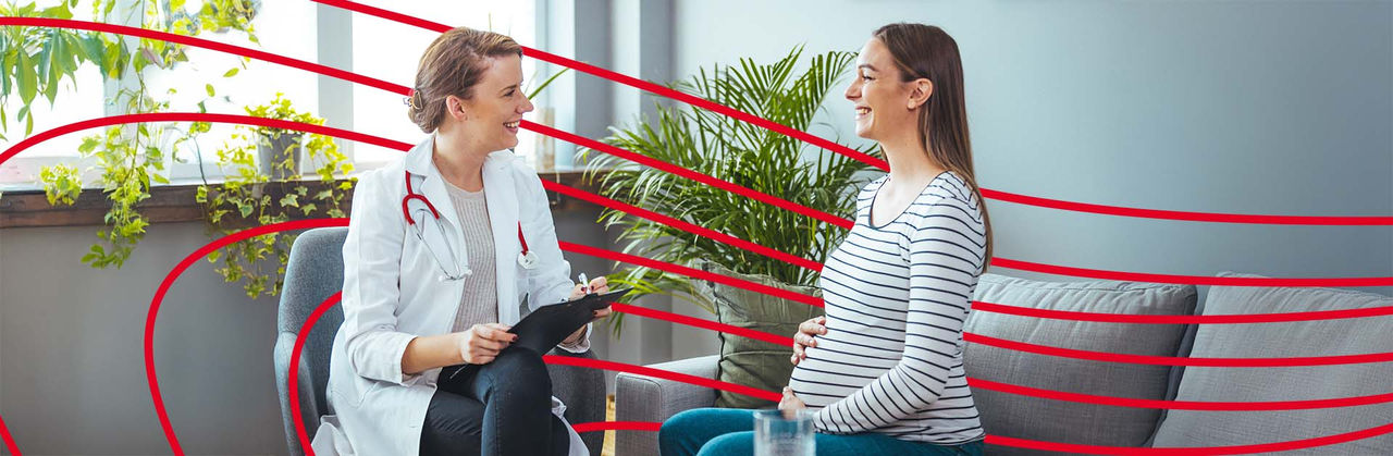 Nurse is a red sweater sitting down on a gray sofa talking with a pregnant patient.