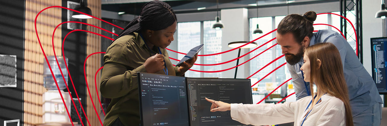 Alt text: A team collaborates around computer monitors displaying code, reviewing technical information and problem-solving together.
