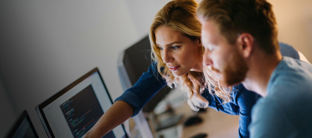 Two IT professionals look at a computer screen while one of them points at it with her right hand.