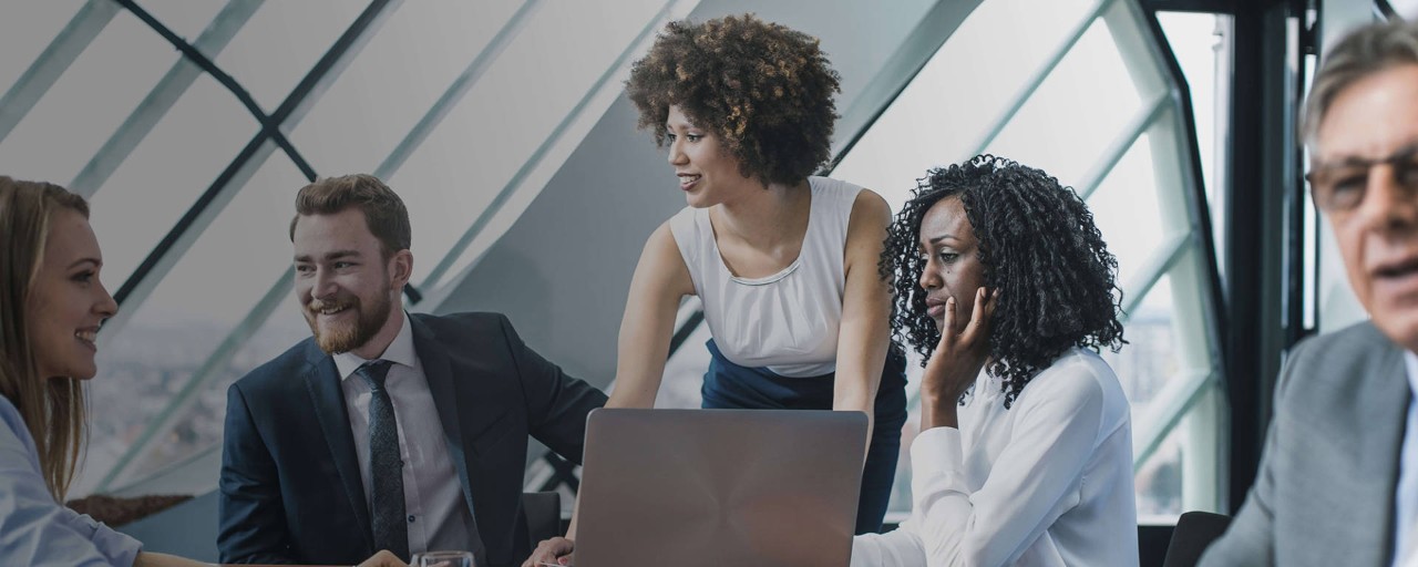 A woman leans over a table during a meeting with four other business professionals, all of whom are sitting around a table that has an open laptop on it.