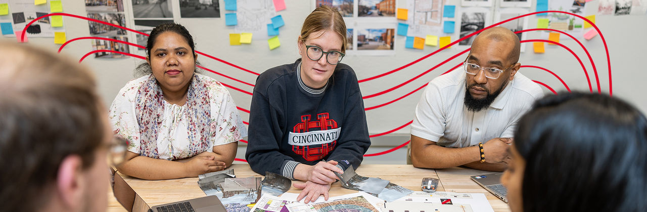 Graduate students collaborating on a city planning project at a table covered with maps and design layouts, with colorful notes and urban development plans on the wall behind them, representing teamwork in the Master of City Planning program.