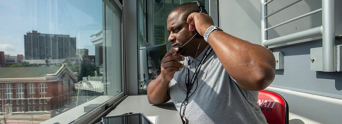 A UC sports administrator in a press box wearing a headset, overlooking campus.