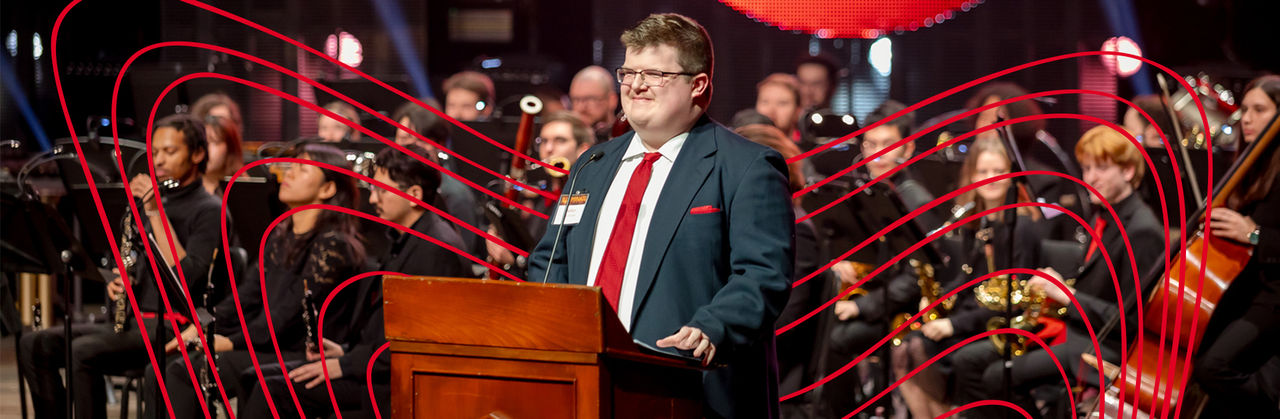 Man wearing suit and glasses standing at podium giving a speech with orchestral band behind him