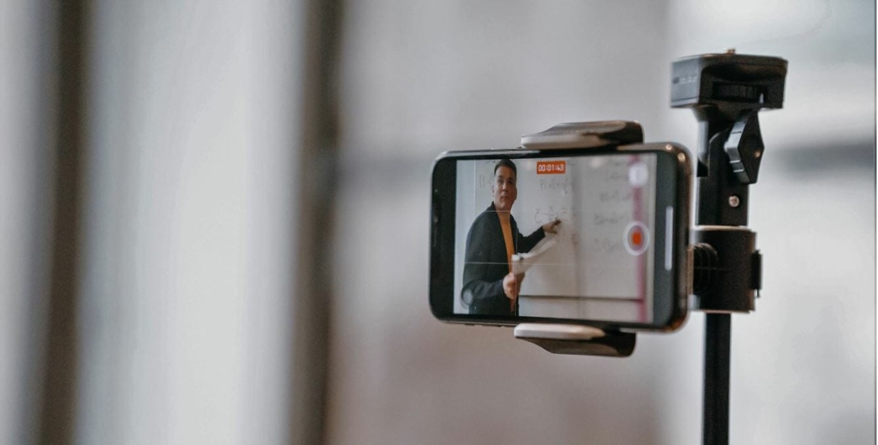 Educator recording an online lesson using a smartphone camera mounted on a tripod, demonstrating concepts on a whiteboard.