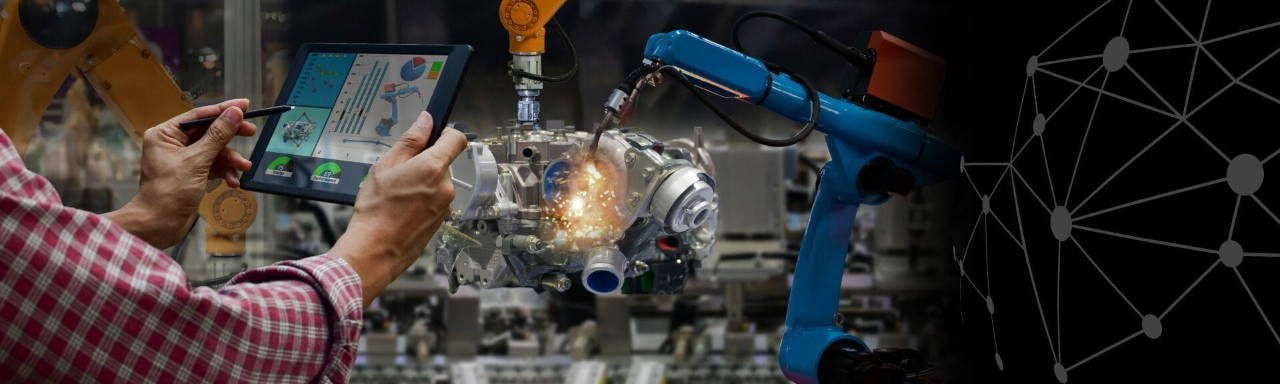 Robotics engineer using a tablet to monitor and control robotic arms performing automated welding in a manufacturing environment.