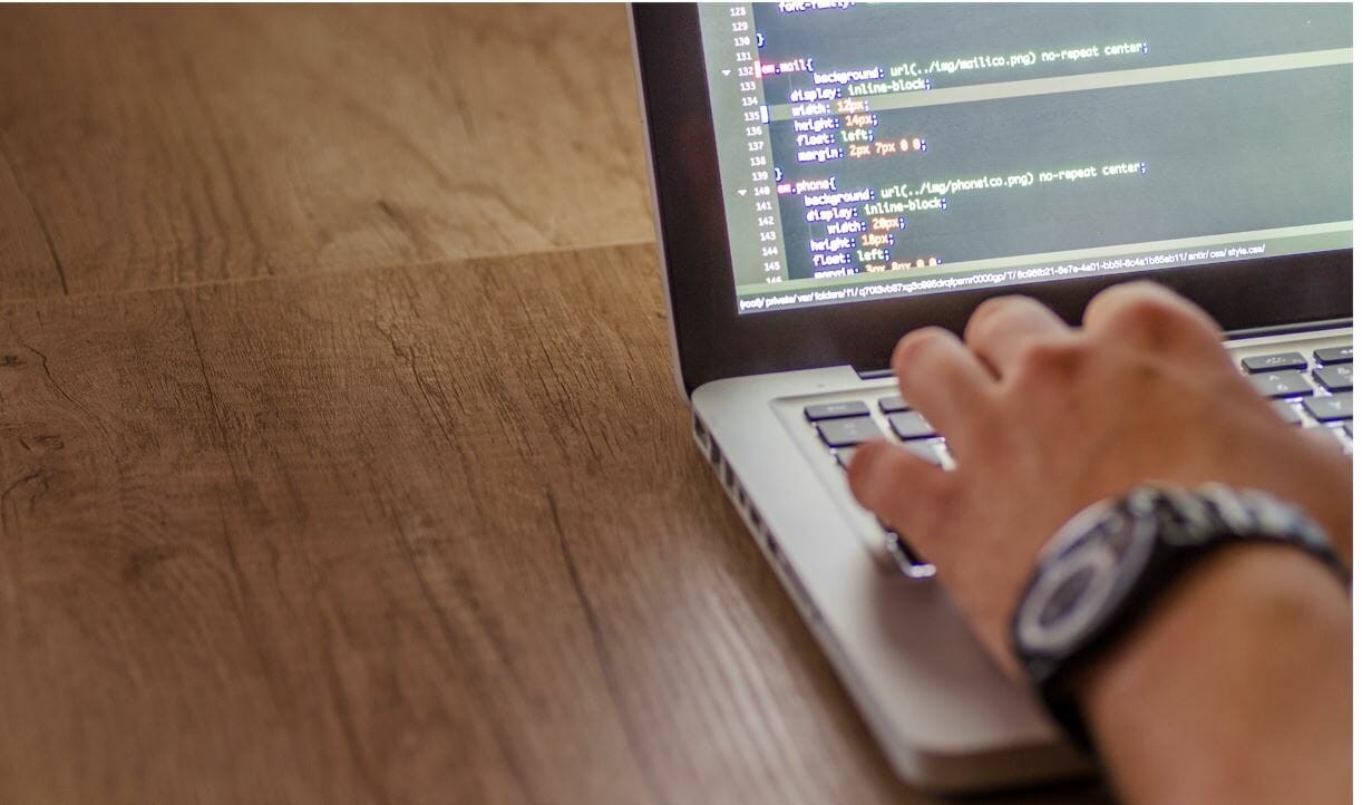 A close-up image of a left hand with a watch on the wrist typing on a laptop with code on the screen.