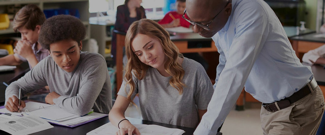 An educator leans over a desk to help a student with her schoolwork while another student sitting next to her looks on.
