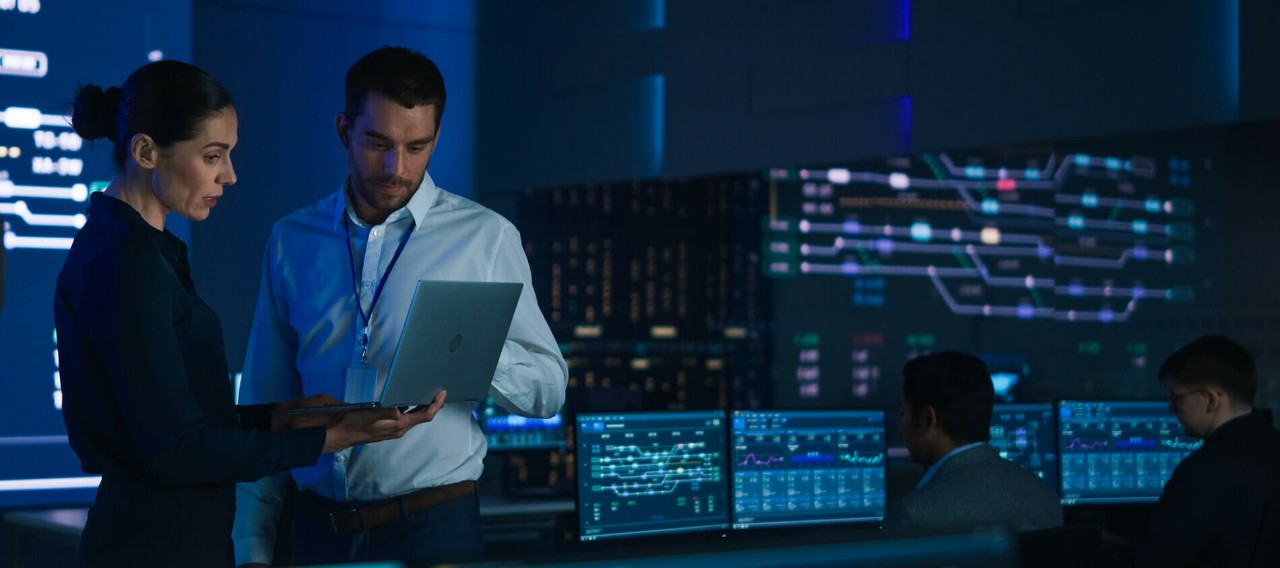 Two cybersecurity professionals stand and look at an open laptop screen while other people are working on dual-monitor computers in the background.