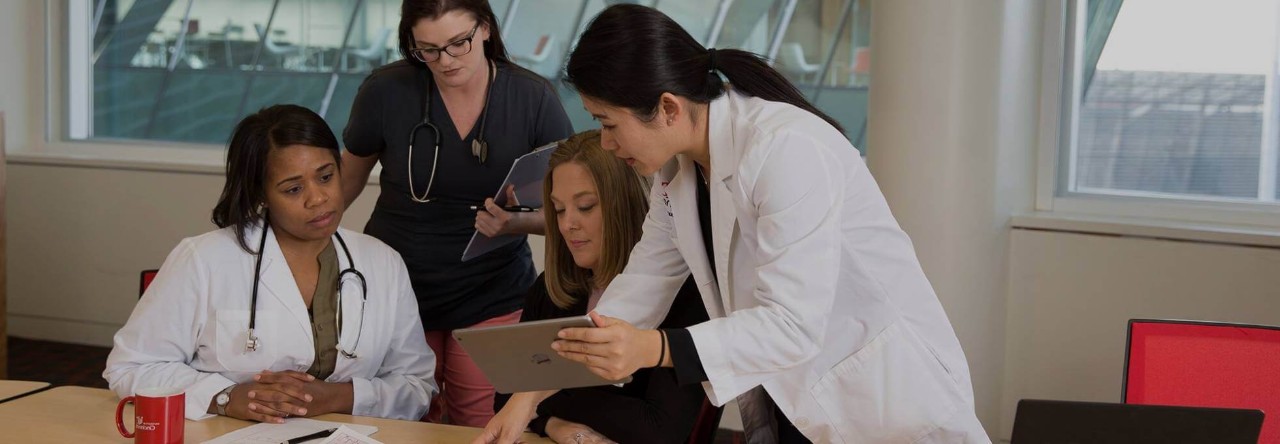 A health data scientist is showing a tablet screen to three coworkers, two of whom are sitting at a table.