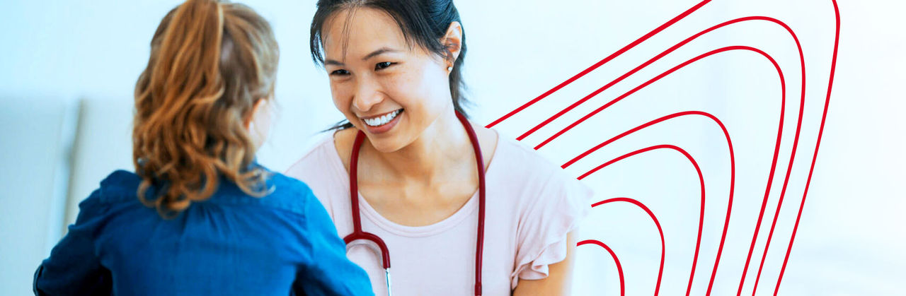 A nurse practitioner wearing a red stethoscope smiles at a child patient.